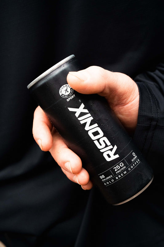 A hand holding a matte black 250ml RISONIX Energy can, front label showing 98 calories, 25g protein and zero sugar.